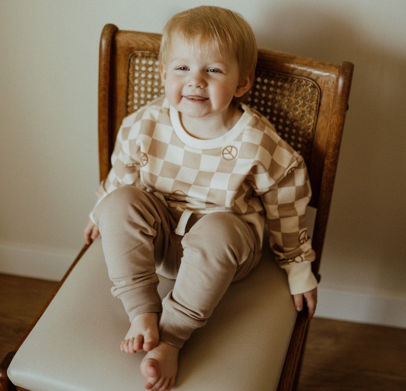 Puede incluir: Un ni&ntilde;o peque&ntilde;o lleva un su&eacute;ter a cuadros marr&oacute;n y blanco con s&iacute;mbolos de paz impresos. El ni&ntilde;o est&aacute; sentado en una silla de madera con un asiento de ca&ntilde;a tejido. El ni&ntilde;o lleva pantalones marrones.