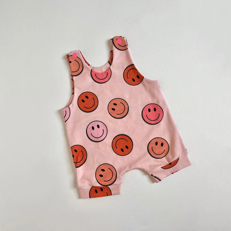 May include: A pink sleeveless baby onesie with a pattern of orange and pink smiley faces.