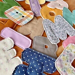 May include: Assortment of fabric letters in various colors and patterns, including floral, polka dot, and plaid designs. Each letter has a scalloped edge. The letters are scattered on a wooden surface.