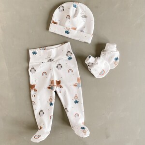 May include: A white baby outfit set with a hat, leggings and mittens. The leggings and mittens feature a woodland animal print with rainbows.