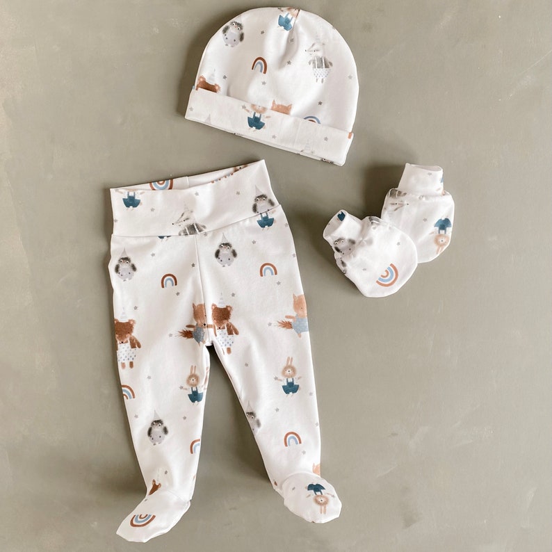 May include: A white baby outfit set with a hat, leggings and mittens. The leggings and mittens feature a woodland animal print with rainbows.