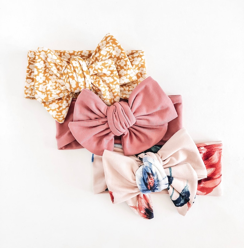 May include: Three fabric headbands with different patterns. The top headband is yellow with white floral print. The middle headband is pink with a large bow. The bottom headband is pink with a floral print and a large bow.