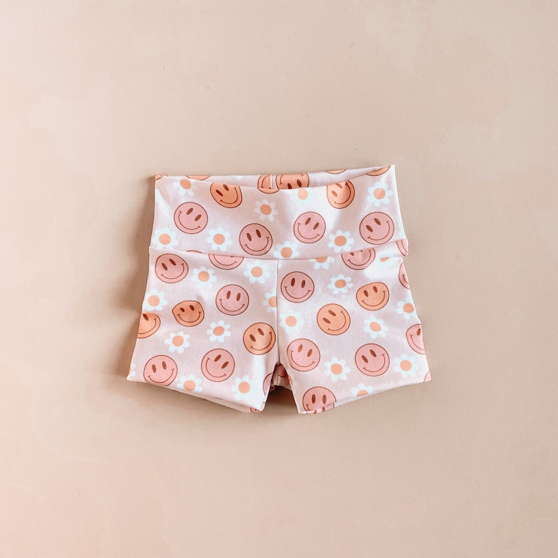 May include: Pink shorts with a pattern of orange and brown smiley faces and white daisies.