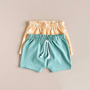 May include: Two pairs of shorts, one in a light blue color and the other in a light yellow color with a white sun pattern. The light blue shorts have a white drawstring.