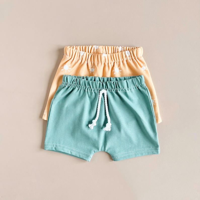 May include: Two pairs of shorts, one in a light blue color and the other in a light yellow color with a white sun pattern. The light blue shorts have a white drawstring.