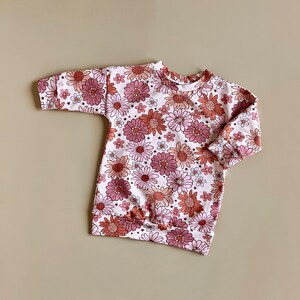 May include: A white long-sleeved sweatshirt with a pink and orange floral print. The sweatshirt has a round neckline and a gathered hem.