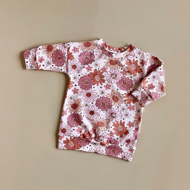 May include: A white long-sleeved sweatshirt with a pink and orange floral print. The sweatshirt has a round neckline and a gathered hem.