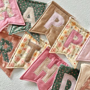 May include: A fabric banner featuring individual pennants in various colours and floral patterns, each displaying a letter. The visible letters spell out parts of the phrase HAPPY BIRTHDAY, with stitched fabric appliqu&eacute;s on each triangular flag.