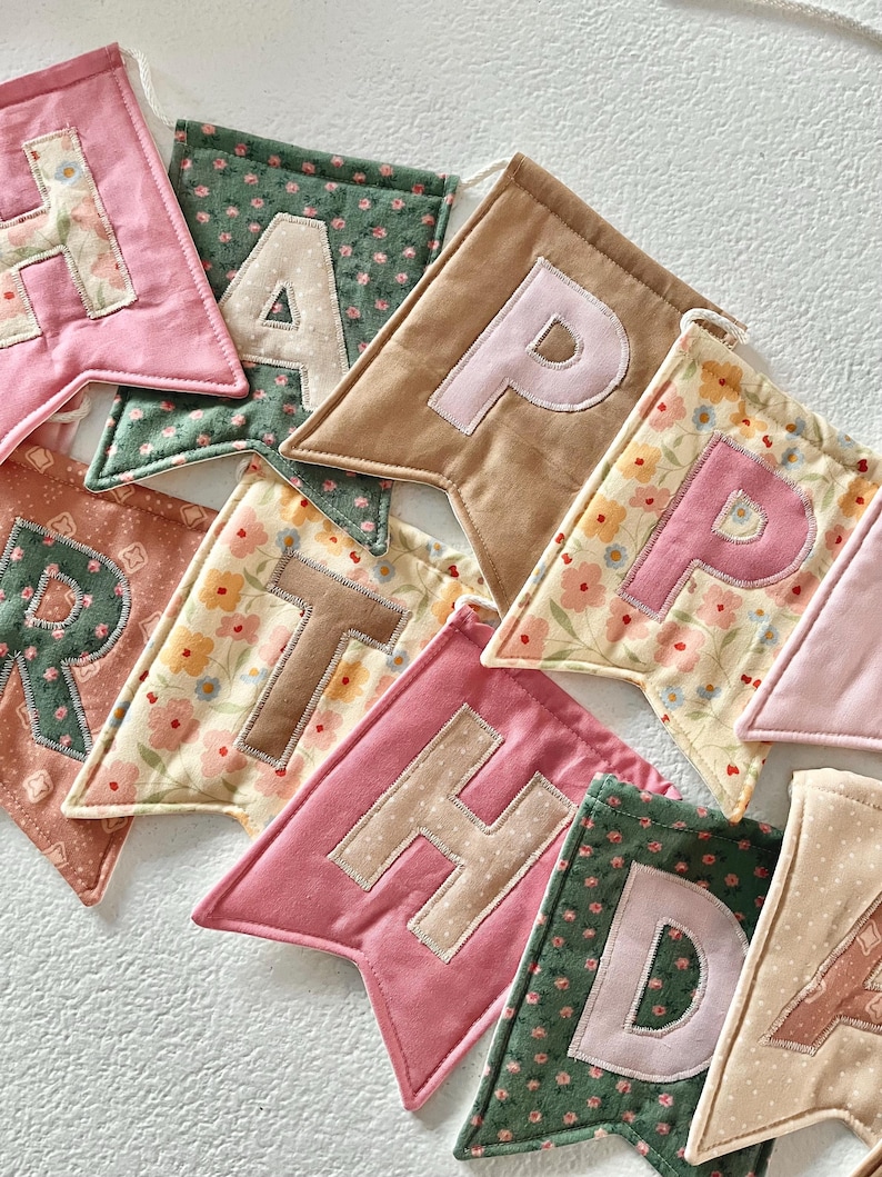 May include: A fabric banner featuring individual pennants in various colours and floral patterns, each displaying a letter. The visible letters spell out parts of the phrase HAPPY BIRTHDAY, with stitched fabric appliqu&eacute;s on each triangular flag.