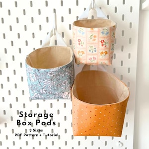 May include: Three fabric storage box pods with white hanging loops. One features a light blue floral pattern, one has a peach grid floral print, and one is orange with white dots. Text reads: Storage Box Pods, 3 Sizes, PDF Pattern + Tutorial.