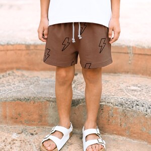 May include: Brown shorts featuring a black lightning bolt pattern and a white drawstring waistband. The shorts are paired with a plain white t-shirt and white double-strap sandals with metallic buckles, creating a casual outfit ensemble.