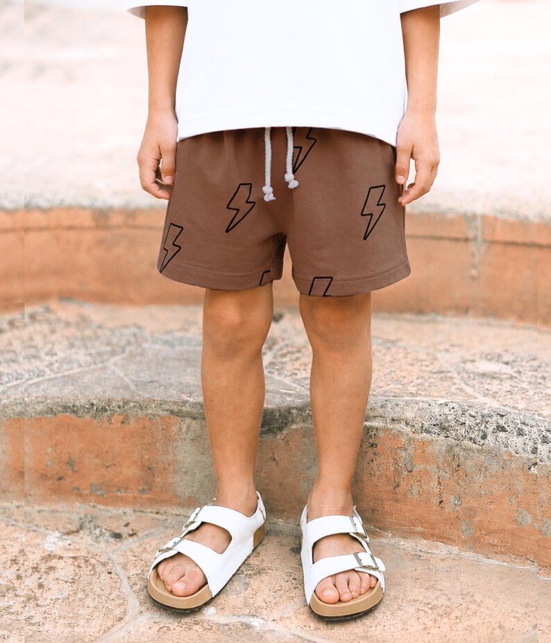May include: Brown shorts featuring a black lightning bolt pattern and a white drawstring waistband. The shorts are paired with a plain white t-shirt and white double-strap sandals with metallic buckles, creating a casual outfit ensemble.