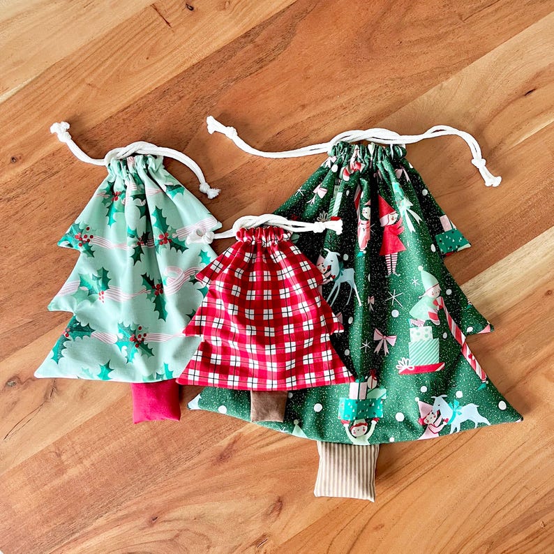 May include: Three Christmas tree-shaped drawstring bags. The bags are made of fabric with festive patterns. One bag is red plaid, another is green with holly, and the largest is green with elves and presents. Each has a white drawstring.