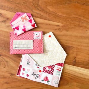 Op de afbeelding: Drie stoffen portemonnees in roze en wit worden getoond op een houten oppervlak. De portemonnees hebben verschillende patronen, waaronder harten, kersen en bloemmotieven. E&eacute;n portemonnee is open en toont een patroon aan de binnenkant en een rode drukknoopsluiting.