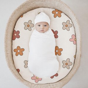 K&ouml;nnte beinhalten: Ein wei&szlig;es, gewickeltes Baby mit einer wei&szlig;en Strickm&uuml;tze liegt in einem geflochtenen Korb mit wei&szlig;em Hintergrund und einem Blumenmuster. Die Blumen sind in Braun-, Orange-, Rosa- und Rott&ouml;nen gehalten.