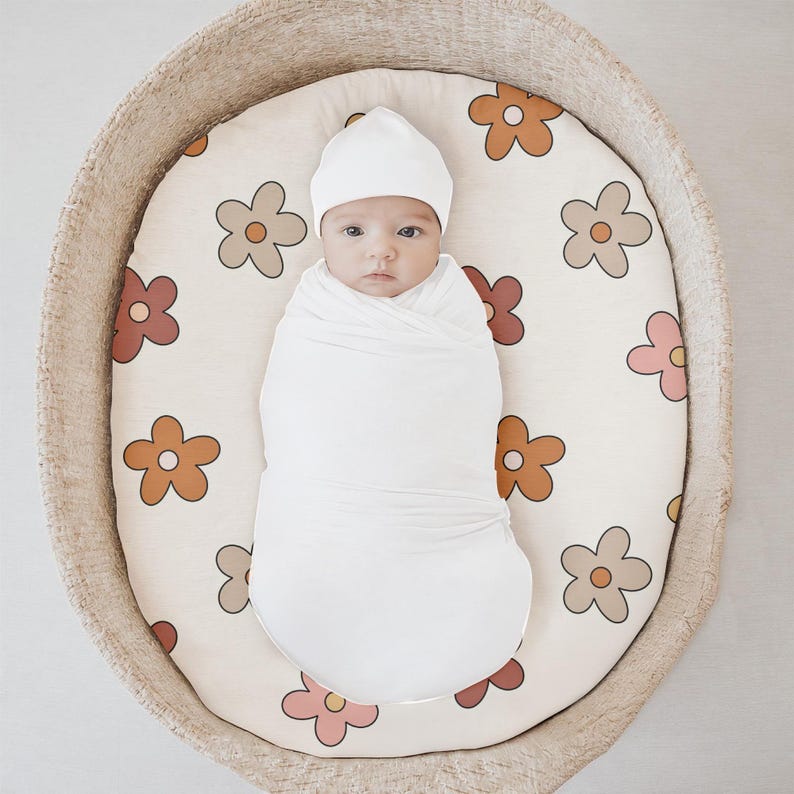 K&ouml;nnte beinhalten: Ein wei&szlig;es, gewickeltes Baby mit einer wei&szlig;en Strickm&uuml;tze liegt in einem geflochtenen Korb mit wei&szlig;em Hintergrund und einem Blumenmuster. Die Blumen sind in Braun-, Orange-, Rosa- und Rott&ouml;nen gehalten.