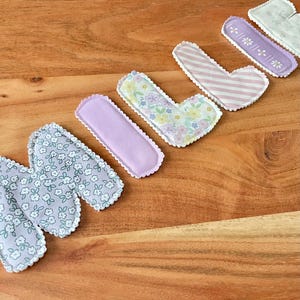 May include: A collection of fabric letters in various patterns and colors, including floral, striped, and solid lavender. Each letter has a scalloped edge, creating a charming, handmade aesthetic. The letters are arranged on a wooden surface.