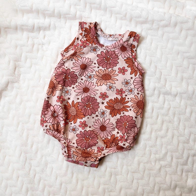 Op de afbeelding: Een roze baby romper met een bloemenprint met roze, oranje en witte bloemen. De romper is mouwloos en heeft een ronde hals.