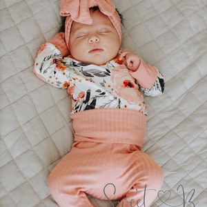 May include: A baby wearing a pink floral print onesie and matching pink pants with a pink bow headband. The baby is sleeping on a white quilted blanket. The image includes the text "Sweet & B Designs".
