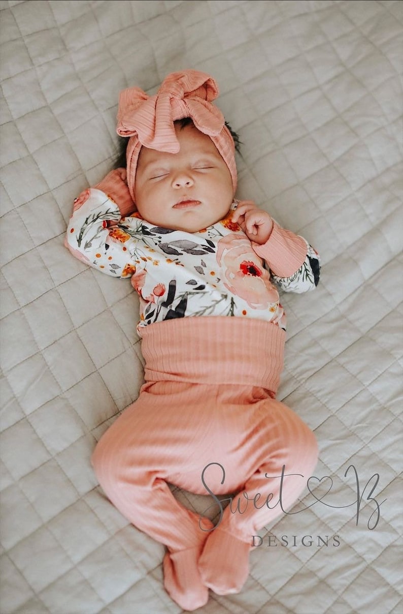 May include: A baby wearing a pink floral print onesie and matching pink pants with a pink bow headband. The baby is sleeping on a white quilted blanket. The image includes the text "Sweet & B Designs".
