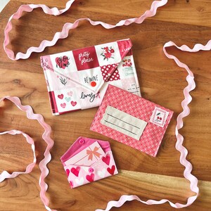 Op de afbeelding: Drie stoffen enveloppen in roze en wit, versierd met harten, bloempatronen en teksten als "You + Me" en "Be Mine". Een roze lint slingert rond de enveloppen op een houten oppervlak. De enveloppen zijn ontworpen met een knoopsluiting.