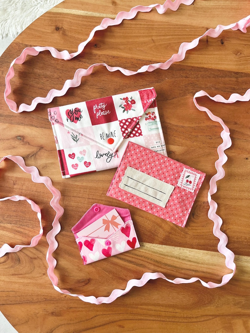 Op de afbeelding: Drie stoffen enveloppen in roze en wit, versierd met harten, bloempatronen en teksten als "You + Me" en "Be Mine". Een roze lint slingert rond de enveloppen op een houten oppervlak. De enveloppen zijn ontworpen met een knoopsluiting.