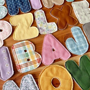 May include: Assortment of colorful fabric letters in various patterns and textures, including polka dots, stripes, floral, and plaid. The letters are in shades of blue, pink, yellow, brown, and green, with a scalloped edge.