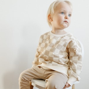 Puede incluir: Un ni&ntilde;o peque&ntilde;o lleva una sudadera de color beige y blanco a cuadros con signos de paz marrones impresos en ella. La sudadera tiene mangas largas y cuello redondo. El ni&ntilde;o tambi&eacute;n lleva pantalones beige.