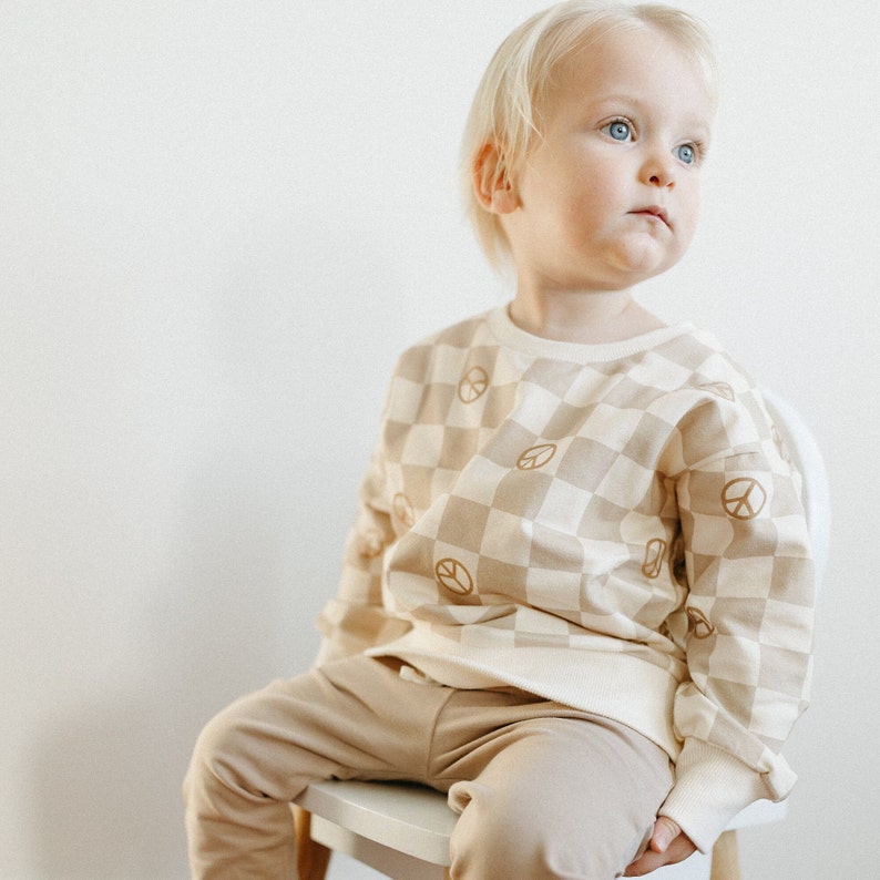 Puede incluir: Un ni&ntilde;o peque&ntilde;o lleva una sudadera de color beige y blanco a cuadros con signos de paz marrones impresos en ella. La sudadera tiene mangas largas y cuello redondo. El ni&ntilde;o tambi&eacute;n lleva pantalones beige.