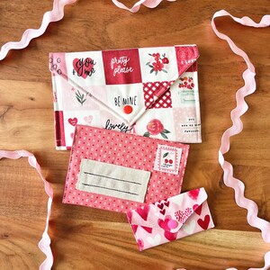 Op de afbeelding: Drie stoffen enveloppen in roze en rood, met Valentijnsdagpatronen en tekst. De grootste envelop heeft een patchwork ontwerp met zinnen als "You + Me" en "Pretty Please". Een roze lint versiert het houten oppervlak.