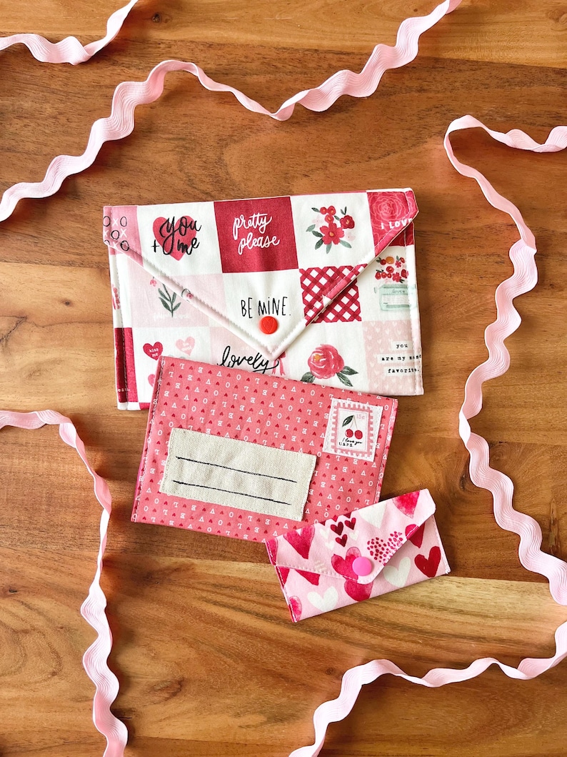 Op de afbeelding: Drie stoffen enveloppen in roze en rood, met Valentijnsdagpatronen en tekst. De grootste envelop heeft een patchwork ontwerp met zinnen als "You + Me" en "Pretty Please". Een roze lint versiert het houten oppervlak.
