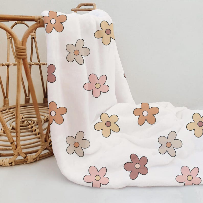 K&ouml;nnte beinhalten: Eine wei&szlig;e Musselin-Wickeldecke mit einem sich wiederholenden Muster aus braunen, rosa und gelben Blumen. Die Decke ist gefaltet und &uuml;ber einen Weidenkorb gelegt.