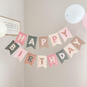 May include: A fabric banner spelling out HAPPY BIRTHDAY in two rows. The pennants are made of various solid and floral patterned fabrics in shades of pink, sage green, and tan. Each letter is appliqued onto the centre of the individual pennant flags.