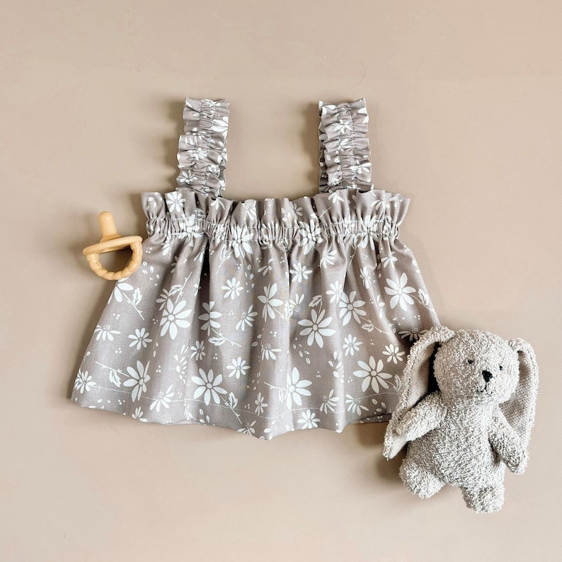 Puede incluir: Un vestido de beb&eacute; gris sin mangas con estampado floral blanco y tirantes con volantes.