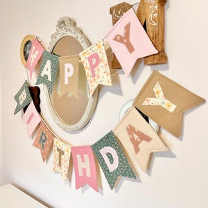 May include: A fabric birthday banner featuring individual pennants in various colours and patterns, including floral prints, solid pink, and tan. Each pennant displays a letter in a contrasting colour, spelling out the phrase HAPPY BIRTHDAY.