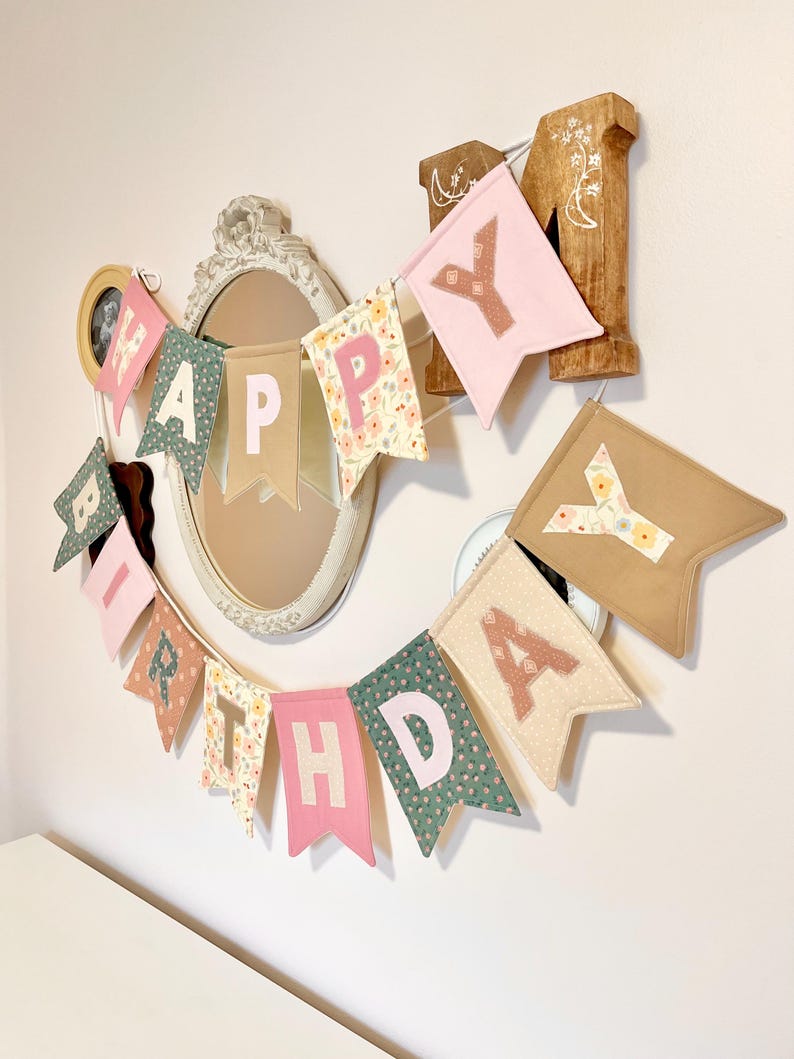 May include: A fabric birthday banner featuring individual pennants in various colours and patterns, including floral prints, solid pink, and tan. Each pennant displays a letter in a contrasting colour, spelling out the phrase HAPPY BIRTHDAY.