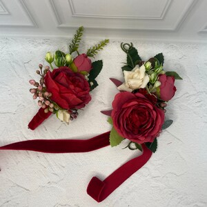 Op de afbeelding: Twee rode en witte bloemcorsages met een bordeauxrood fluwelen lint. De corsages zijn gemaakt van zijden bloemen en groen.