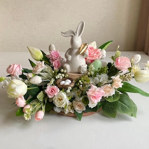Könnte beinhalten: Eine weiße Keramik-Hasenfigur, die eine Karotte hält, sitzt in der Mitte eines Blumenarrangements. Das Arrangement besteht aus rosa, weißen und grünen Blumen sowie einem Nest mit weißen Eiern.
