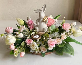 Peut inclure: Une figurine de lapin en céramique blanche tenant une carotte est placée au centre d'un arrangement floral. L'arrangement comprend des fleurs roses, blanches et vertes, ainsi qu'un nid avec des œufs blancs.