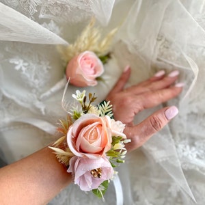 May include: A pink and white floral wrist corsage featuring a single large rose and smaller roses, accented with greenery and dried grasses.