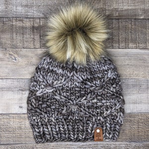 Puede incluir: Un gorro de punto gris y negro con un gran pompón de piel sintética en la parte superior. El gorro tiene una etiqueta de cuero marrón con el texto "Roots & Cedar" en ella.