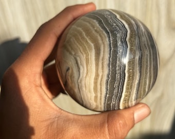 May include: A polished, spherical stone with bands of tan, gray, and white. The orb is held in a hand, showcasing its smooth surface and natural color variations. The stone's banded pattern creates a unique, earthy aesthetic.