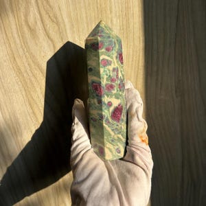 Può includere: Un obelisco di pietra preziosa verde e rossa. L'obelisco è tenuto in una mano guantata e presenta un mix di colori verdi, rossi e crema. La pietra preziosa ha una superficie lucida e liscia e una parte superiore appuntita. Lo sfondo è una superficie di legno.