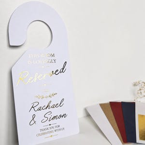 Könnte beinhalten: Ein weißer Türhänger mit goldener Folienprägung, der den Text THIS ROOM IS LOVINGLY Reserved FOR Rachael & Simon THANK YOU FOR CELEBRATING WITH US trägt. Der Hänger hat eine Hakenform und liegt neben einem Set aus farbigen Briefumschlägen.