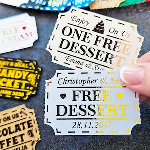 Könnte beinhalten: Eine Sammlung rechteckiger Papiertickets mit gewelltem Rand, verziert mit metallischer Folienprägung. Ein weißes Ticket zeigt "Enjoy ONE FREE DESSERT On Us" mit einem Tortensymbol, ein weiteres "Christopher & Amy FREE DESSERT" mit Herzsymbolen.