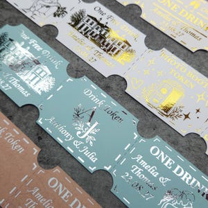 Puede incluir: Una colección de boletos de papel rectangulares con bordes perforados, que presentan diseños de lámina metálica en oro y plata. Los boletos muestran textos como One Free Drink, Drink Token y Photo Booth Token, junto con ilustraciones de un edificio, una cabina de fotos y nombres con fechas.