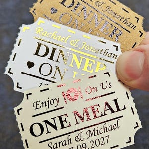 Könnte beinhalten: Drei rechteckige Papiertickets mit gewelltem Rand. Das oberste Ticket ist hellbraun mit schwarzem Text Rachael & Jonathan DINNER ON US. Das mittlere ist weiß mit goldener Schrift Rachael & Jonathan DINNER ON US. Das untere ist cremefarben mit roter Schrift Enjoy On Us ONE MEAL Sarah & Michael 07.09.2027.