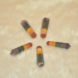 May include: Five seven chakra crystal wands, each with a different color band, arranged in a star shape on a white fuzzy surface.