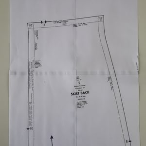 Op de afbeelding: Een zwart-wit naaipatroon voor de achterkant van een rok. Het patroon bevat instructies voor het knippen, naaien en plaatsen van de stof. Het patroon is voor maat 5 en is gemarkeerd met "SKIRT BACK".