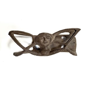 May include: A carved wooden wall hanging featuring a stylized human figure with outstretched arms and a serene facial expression. The dark brown wood has intricate details, including textured patterns along the arms and head. The piece has two holes for mounting.
